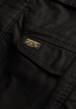 Bolsa de tela negra con una etiqueta cosida en la tapa que dice "Superdry Utility Wear 360-366/054 MFG 'CO'" en negro y amarillo.