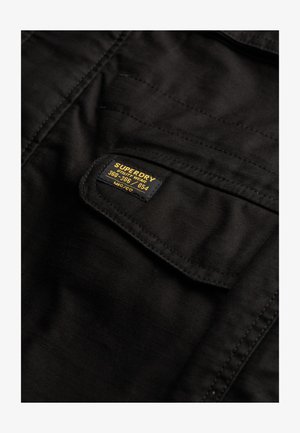 Zwarte stoffen zak met een zwart en geel label "Superdry Utility Wear 360-366/054 MFG 'CO'" gestikt op de flap.
