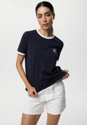 Kobieta z ciemnymi włosami w kucyku, ubrana w granatowy t-shirt z białym wykończeniem i białe szorty, trzymająca dolną część t-shirtu.