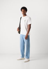 Witte katoenen t-shirt met een kleine grafische print op de linkerborst, gecombineerd met lichtblauwe jeans en witte sneakers. Het model houdt een groene tas vast.