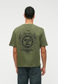 Õlivaheline t-särk, mis on valmistatud puuvillast, musta graafilise logoga, millel on laurolilled tagaküljel. Lühikesed varrukad ja lõdvestatud istuvus.