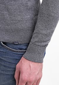 Gros plan sur la main d'une personne reposant à ses côtés, portant un pull gris et un jean bleu avec une petite étiquette indiquant "1985."