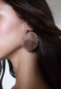 Orecchini a pompom in pelliccia sintetica marrone su un gancio dorato. Texture morbida, forma rotonda, posizionati vicino al collo di una donna con capelli scuri fluenti.