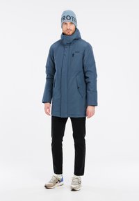 Parka isolante bleue avec un col haut, des poches latérales et une poche zippée sur la poitrine. Associée à un jean noir et des baskets multicolores.