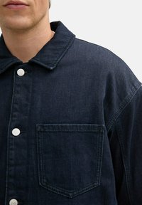 Czarna dżinsowa kurtka z wyrazistym kołnierzem, białymi guzikami oraz jedną kieszenią na piersi w charakterystycznym prostokątnym kształcie.