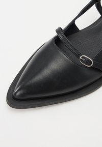 Schwarzer Lederschuh mit spitzer Zehenform, ausgestattet mit einem Riemen über dem Fußrücken und einer silbernen Schnalle. Texturiertes Innenfutter und Sohle.