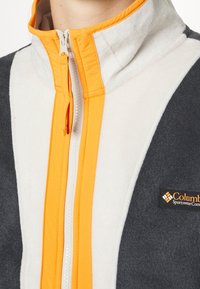 Lähikuva harmaasta ja valkoisesta fleece-takista, jossa on oranssi vetoketju ja Columbia Sportswear Companyn logo-ompelu rintataskussa.