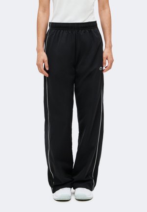 Personne portant un pantalon de survêtement noir ample avec des bandes blanches sur les côtés, des baskets blanches et un haut blanc, debout avec les bras détendus le long du corps.