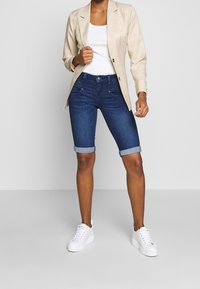 Kvinna iklädd uppvikta mörkblå denimshorts, vit linne, beige rutigt kostymjacka och vita plattformssneakers, stående mot en enhetlig bakgrund.