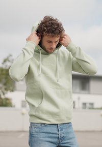 Lichtgroene hoodie met een voorzak, trekkoorden en geribbelde manchetten; gedragen met blauwe jeans tegen een vervaagde stedelijke achtergrond.