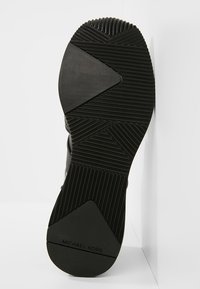 Semelle de chaussure noire avec des motifs géométriques de la bande de roulement et la marque "MICHAEL KORS" sur la zone du talon sur fond blanc.