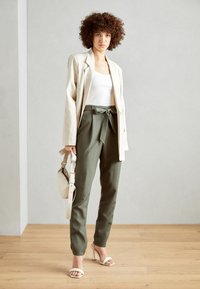 Hellbeige Leinenblazer über einem weißen Tanktop, kombiniert mit olivgrünen, hochgeschnittenen Hosen und weißen offenen Schuhen. Kleine Tasche inklusive.