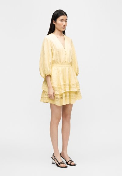 Νεαρή γυναίκα με ανοιχτό κίτρινο φόρεμα με στρώσεις και φουσκωτά μανίκια, φορόντας μαύρα πέδιλα με λουράκια και τακούνια, στέκεται μπροστά σε λευκό φόντο.