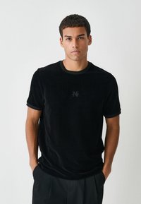 Schwarzes Cord-T-Shirt mit kurzen Ärmeln, gerippter Textur, rundem Halsausschnitt und subtil eingesticktem Logo auf der Brust. Passender dunkler Besatz.