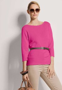 Frau in einem leuchtend pinken Langarmoberteil mit schwarzem Gürtel, beigen Hosen, großen Sonnenbrillen, die eine geflochtene braune Handtasche und hölzerne Armbänder hält.