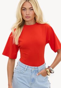 Femme aux longs cheveux blonds portant un haut moulant rouge à manches courtes et un jean taille haute bleu clair avec des boutons métalliques et des bracelets.
