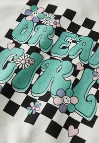 Bomullsdesign med teksten "DREAM GIRL" i blågrønn farge, omgitt av blomster og hjerter, på en svart-hvit sjakkbrettbakgrunn.