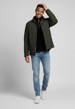 Chaqueta acolchada verde oscura con cuello alto y cremallera, llevada sobre un suéter negro, combinada con jeans azul claro y zapatillas blancas.