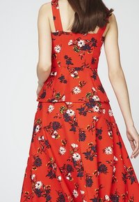 Czerwona sukienka peplum z kwiatowym wzorem, z dopasowaną górą, szerokimi ramiączkami i rozkloszowaną spódnicą. Materiał ma teksturowany wzór z białymi i granatowymi kwiatami.