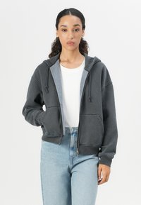 Gray zip-up hoodie with a front pocket, drawstring hood, and ribbed cuffs, layered over a white shirt and paired with light blue jeans.