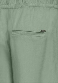Close-up van lichtgroene stofbroek met een elastische tailleband en een smalle achterzak met een klein metalen "CECIL" label.
