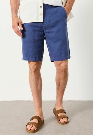 Mann trägt blaue knielange Shorts, ein weißes Hemd und braune Sandalen mit Schnalle, steht mit einer Hand in der Tasche auf einem grauen Boden.