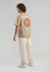 Valkoinen puuvillapaita, jossa on oranssi kukkakuva ja "CALIFORNIA" -teksti selkäpuolella, yhdistettynä vaaleisiin cargo-housuihin ja valkoisiin lenkk kenkiin.