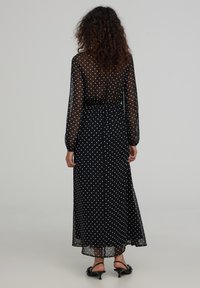 Czarna maxi sukienka z długimi przezroczystymi rękawami, ozdobiona wzorem w kropki. Talie podkreśla skośny krój, a materiał ma lejący, lekki charakter.