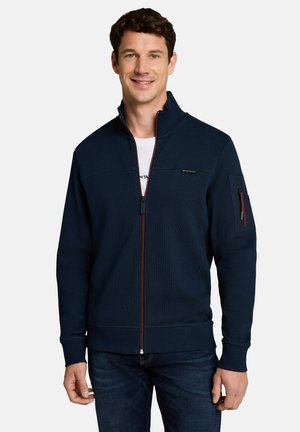 Uomo che indossa una giacca blu navy con zip e dettagli rossi sulla zip e una camicia bianca sotto, in piedi davanti a uno sfondo chiaro e uniforme.