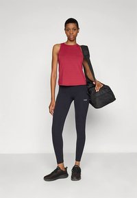 Top atletico senza maniche color bordeaux, leggings neri e sneakers neri. La modella tiene una borsa da palestra nera e si trova di fronte a uno sfondo semplice.