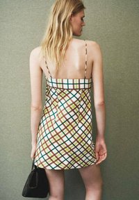 Mujer con cabello largo y rubio que lleva un vestido mini sin mangas y sin espalda de cuadros, sosteniendo un pequeño bolso negro frente a un fondo verde liso.