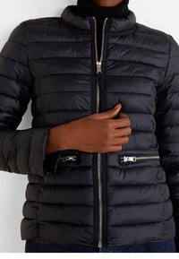 Czarna kurtka puchowa z zapięciem na zamek błyskawiczny z przodu, stójką i dwiema kieszeniami zapinanymi na zamek. Posiada poziome przeszycia i elastyczne mankiety.