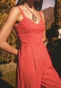 Jumpsuit rosso senza maniche con un busto aderente, tessuto texturizzato caratterizzato da ricami floreali e pantaloni larghi. Completato con una collana audace.