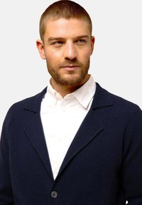 Marinblå ullcardigan med sjalkrage, enkel knappstängning, texturerad tyg, bärs över en vit skjorta med krage.