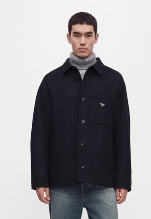 Homme portant une veste noire boutonnée sur un pull gris à col roulé côtelé et un jean bleu, debout devant un fond blanc uni.