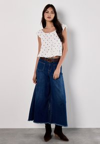 Top bianco plissettato con maniche corte a balza; pantaloni in denim blu a gamba larga con tasche e orlo grezzo; cintura marrone; stivali ankle marroni.