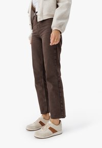 Isik kannab tumeda pruuni kõrge vöökohtadega sirgeid pantalone, beeže tossusid pruunide triipudega, beeži jakk ning kuldringi paremas käes.