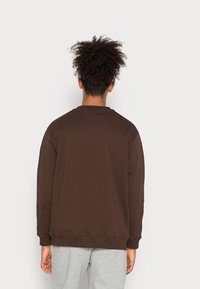 Sudadera marrón con cuello redondo y mangas largas, con un corte holgado. El material parece suave; combinada con pantalones deportivos gris claro.