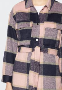 Kobieta nosząca zapinany na pasek płaszcz w kratę w kolorze różowym, czarnym i beżowym, z kieszeniami na klatce piersiowej i srebrnymi zatrzaskami na białej bluzce.