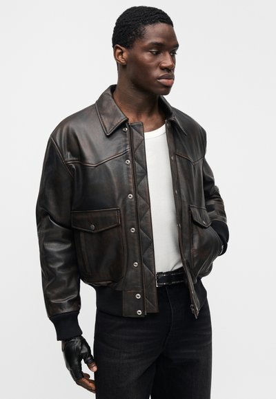 Καφέ δερμάτινο bomber μπουφάν με σχεδιασμό καπιτονέ, κουμπιά με πατιλέτα και πλαϊνές τσέπες. Διαθέτει ριμπ μανσέτες και γιακά. Φοράται πάνω από λευκό πουκάμισο.