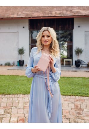 Light blue dress with long sleeves and a tie belt, holding a small, soft pink crossbody bag with a logo. Outdoor setting with green grass.