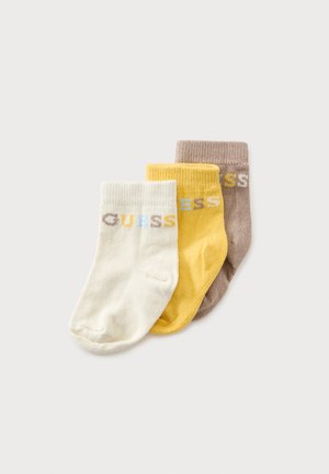 Drei gefaltete Babysocken in Creme, Gelb und Hellbraun, jeweils mit dem Wort "GUESS" in mehrfarbigen Buchstaben nahe dem Bündchen gestrickt.