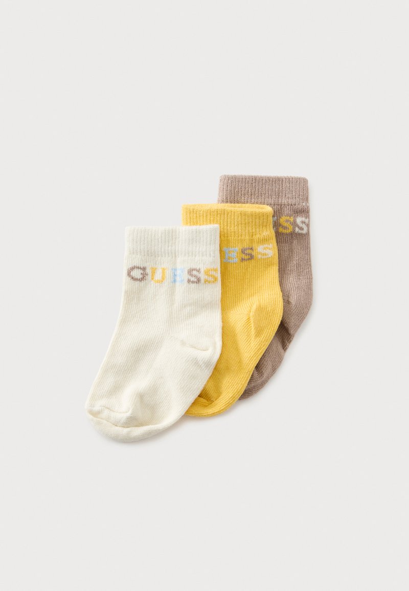 Trois chaussettes bébé pliées en crème, jaune et brun clair, chacune avec le mot "GUESS" tricoté près du bord en lettres multicolores.