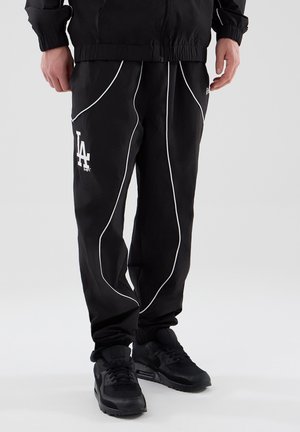 Person iført sorte bukser med LA-logo og hvide striber samt sorte sneakers stående mod en lys baggrund.