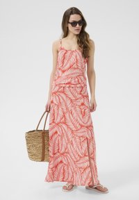 Robe maxi à motifs de feuilles rouges avec des fines bretelles, une taille cintrée et une fente sur le côté. Assortie à un sac en paille tressé et des sandales plates.