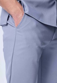 Un adulte portant un pantalon bleu clair et une chemise assortie, avec une main glissée dans la poche latérale.