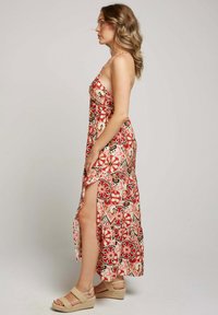 Robe maxi à motif floral rouge et crème avec une fente sur le côté, de fines bretelles et une coupe décontractée ; associée à des sandales compensées.