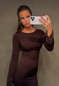 Femme aux longs cheveux bruns portant une robe marron foncé ajustée, prenant un selfie dans un miroir avec un smartphone blanc, portant des boucles d'oreilles créoles et un bracelet.