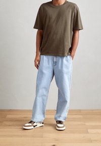 Olive green cotton t-shirt, relaxed fit; light blue denim trousers, loose style; brown and white sneakers with lateral stripes.