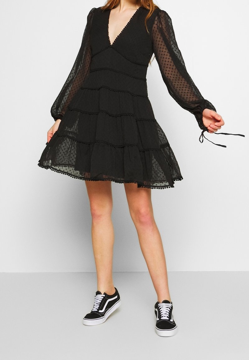 Femme portant une robe noire à pois à manches longues avec une jupe à étages et des baskets noires et blanches, debout devant un fond uni.